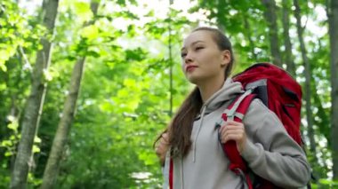Young woman wearing red backpack goes hiking smiling in huge dense forest. Brown-haired lady enjoys hiking uniting with wild nature in morning slow motion