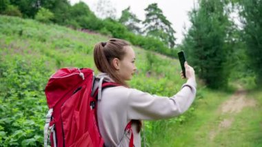 Brown-haired woman travel blogger makes selfies on smartphone. Happy young hiker wearing red backpack records video of herself against flower meadow