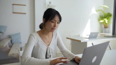 Focused Asian woman types report on laptop keyboard sitting at desk in modern office. Young employee works online using electronic device closeup