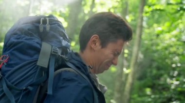 Black-haired young man uses mosquito spray on face and whole body grimacing. Guy holding blue backpack hates insects hiking in dense forest closeup slow motion