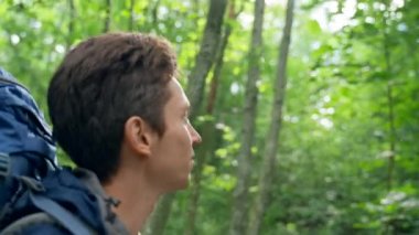 Black-haired young man enjoys hiking uniting with nature. Guy with blue backpack walks smelling fresh fragrances of green dense forest closeup