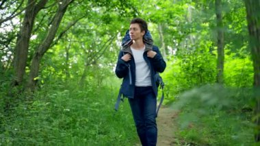 Young man with blue backpack walks in thick forest smiling. Black-haired guy wearing blue jacket and white shirt enjoys uniting with wild nature slow motion