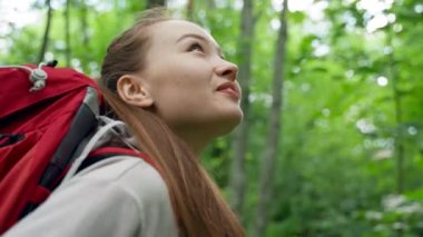 Brown-haired young woman walks in dense forest enjoying hiking. Sun rays break through thick branches. Lady hiker walks with backpack smiling closeup
