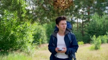 Black-haired guy hiker walks on meadow smiling and looking at phone for finding destination. Young man with blue backpack prefers hiking in forest