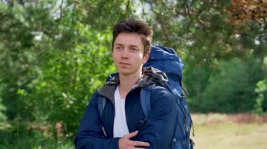 Young man hiker stands on green forest glade looking around for footpath for hiking. Black-haired tourist enjoys walking uniting with forest nature