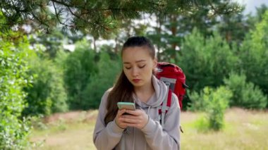 Brown-haired woman types notes on smartphone about picturesque forest walking past green trees. Young lady enjoys hiking in huge forest