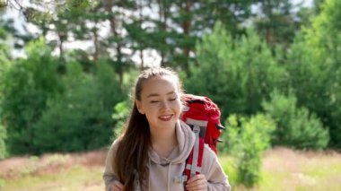 Brown-haired woman walks on meadow smiling in sunny morning. Young female hiker wearing grey trousers and sweatshirt enjoys hiking on forest glade