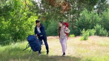 Young people hikers put bright backpacks on grass to rest against trees. Couple of tourists puts things on grass from backpacks for camping on forest glade