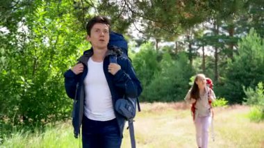 Couple of young campers enjoys hiking together. Girlfriend and boyfriend hikers with backpacks walk discussing things and smile on forest glade
