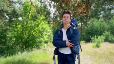 Young man hiker wearing blue jacket and white shirt stands in forest looking thoughtfully. Black-haired guy enjoys hiking on forest glade on sunny day