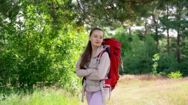 Happy young woman goes hiking in forest with red backpack. Brown-haired tourist enjoys walking and hiking on forest glade smiling with crossed hands