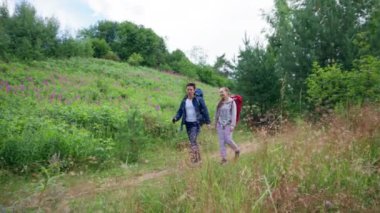 Couple of young tourists walks on footpath trying to find perfect spot for camping. Man and woman enjoy hiking walking past meadow with smile slow motion