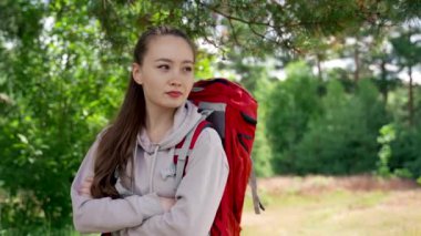 Young woman hiker looks with happy and excited expression on face. Brown-haired tourist with red backpack enjoys hiking on lively green forest glade
