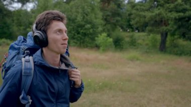 Black-haired man hiker looks around forest listening to music. Young guy wearing headphones with blue backpack enjoys hiking alone in forest closeup