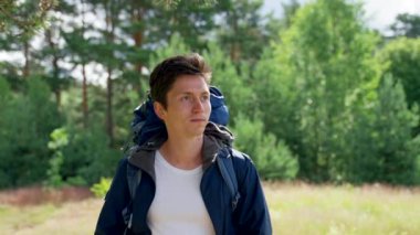 Black-haired guy hiker with blue backpack enjoys walking in forest. Young man discovers awesome landscape hiking in forest with smile on sunny day