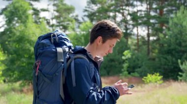 Young man with blue backpack looks on smartphone to find perfect spot for camping in forest. Black-haired person enjoys hiking on green meadow