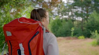 Young woman with red backpack walks on glade searching for perfect destination on sunny day. Brown-haired lady goes hiking in forest backside view slow motion