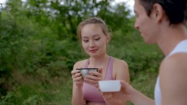 Couple of happy tourists drinks hot beverage in fresh air. Young people sit holding big mugs smiling to each other against blurry green trees closeup