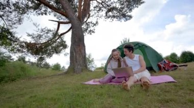 Couple of excited tourists enjoys watching video on laptop waving heads against camp tent. Young hikers like to spend vacation in campground on forest meadow