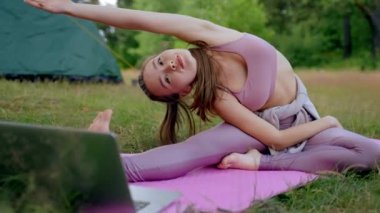 Brown-haired young woman enjoys practicing pilates on mat by camp tent. Lady tourist wearing pink top and leggings does yoga watching video on own laptop