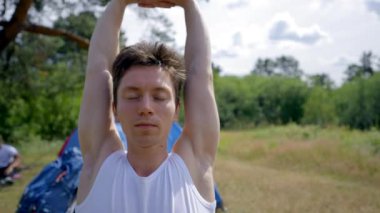 Young man does workout on black mat stretching hands and back. Black-haired camper enjoys healthy vacation on forest meadow with friends on sunny day
