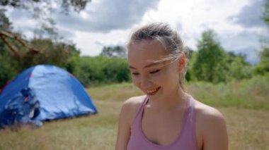 Young woman enjoys working in fresh air by tent on meadow. Brown-haired employee sits on mat typing and searching information via digital device smiling closeup