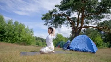 Black-haired hiker practices yoga on mat put on grass by camp tents on meadow. Sunlight illuminates sportsman strong body. Young man enjoys weekend in forest