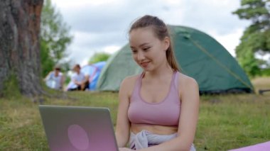 Brown-haired woman works on laptop smiling. Young lady wearing pink top and leggings enjoys working freelancer in free time uniting with nature against camp