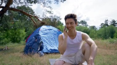 Black-haired man sits on mat in campsite talking on smartphone. Young guy talks smiling and laughing. Sunlight illuminates man strong body near camp tent
