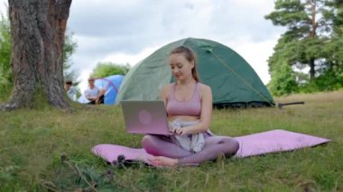 Young woman types useful information for subscribers with smile. Brown-haired manager enjoys freelance in pastime. Lady sits on mat against camp tent