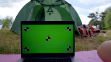 Couple watches laptop with chromakey green screen together on grass against green camp tent. Man and woman enjoy spending time together on vacation in campground closeup