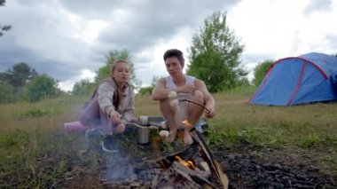 Couple of tourists enjoys cooking marshmallows on campfire against blue camp tent. Happy man and woman discuss things about vacation in dull evening