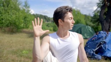 Young man practices yoga against bright blue and green tents. Black-haired guy enjoys healthy lifestyle. Sunlight illuminates hiker face on meadow closeup