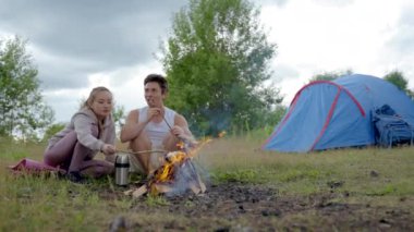 Young couple sits on mat near campfire frying marshmallows. Handsome man smiles and laughs looking at woman burnt down marshmallow on wooden stick