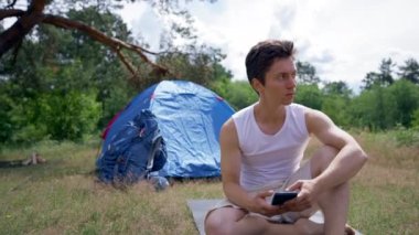 Young man looks on digital map on black smartphone and tries to find and share location. Handsome hiker looks around sitting against blue tent