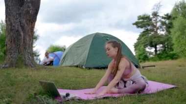 Young woman stretches legs on pink mat. Brown-haired camper puts effort in practicing yoga in fresh air. Lady hiker enjoys vacation on nature in campground