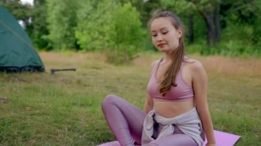 Young female camper stretches body on pink sport mat in fresh air. Brown-haired woman with long ponytail enjoys practicing yoga by tent against blurry green trees