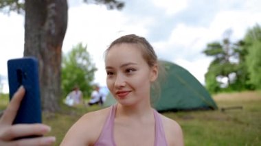 Young woman makes selfie grimacing on phone camera. Brown-haired lady shows vacation in meadow nature against campsite. Camper demonstrates healthy life closeup