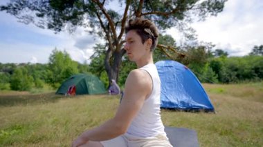 Black-haired hiker does yoga on mat put on grass against forest campsite. Young man follows necessary rules of yoga watching online video course closeup