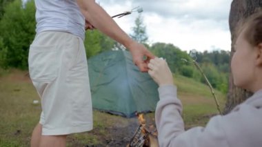 Man and woman cook marshmallows on campfire on meadow against green tent. Hot summer weather changes to cold and dull. Guy gives marshmallow to lady closeup