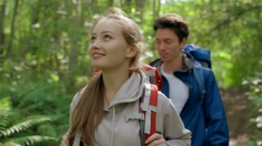 Young girlfriend holding backpack enjoys hiking with boyfriend in deep forest. Happy woman with ponytail walks examining landscape smiling closeup