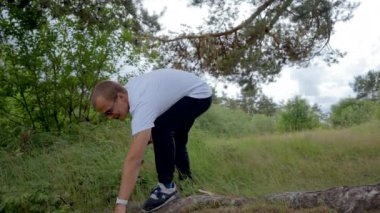 Young man aggressively chops wood on forest meadow. Bald guy in sunglasses wearing white t-shirt and black jeans prepares woods for campfire for cooking food