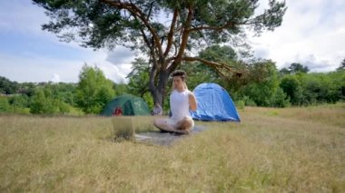 Young man practices yoga sitting on black mat on forest glade meadow. Black-haired hiker enjoys doing workout against picturesque forest landscape