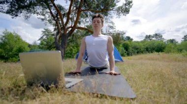 Black-haired camper does workout on black mat looking in laptop. Young man finds online video course of yoga and decides to try exercises near camp tent against sky