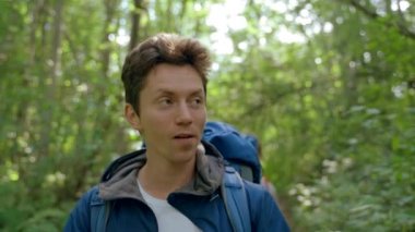 Boyfriend enjoys hiking with girlfriend in deep forest. Happy tourist wearing blue jacket and white shirt walks looking around smiling closeup. Woman follows man