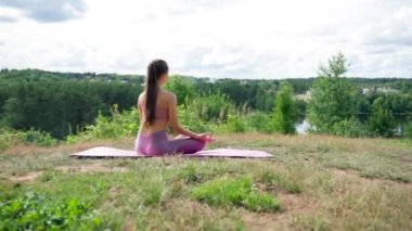 Brunette woman sits on mat meditating in Lotus pose under cloudy sky. Lady with ponytail in top enjoys peaceful day breathing fresh air on hilly river bank