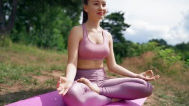 Young smiling woman meditates on mat in Lotus pose on grassy meadow in sunny weather. Tranquil lady in sports suit sits against trees breathing fresh air