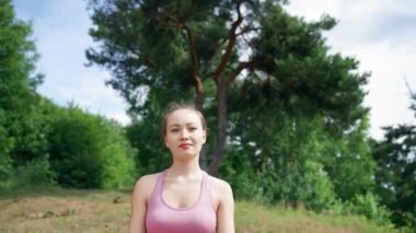 Young woman sits on mat meditating in Lotus pose on grassy meadow. Lady in pink sports suit unites with nature against high green trees closeup