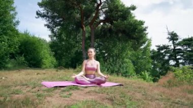 Young brunette meditates in Lotus pose with closed eyes on grassy meadow. Lady in pink sports suit sits smiling on mat against trees breathing fresh air