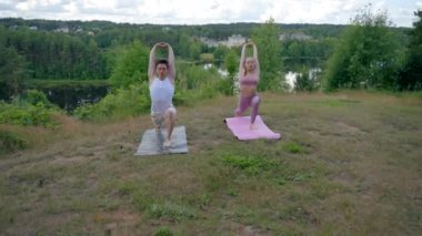 Young man and woman friends practice yoga Warrior pose on mats on steep river bank together. People concentrate on asana enjoying moment on high hill